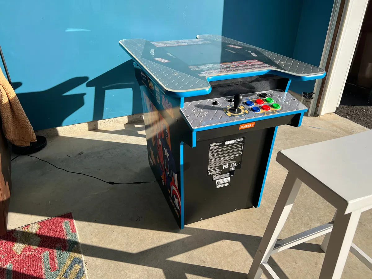 A vibrant interior shot of the garage game room, featuring an arcade machine with colorful buttons set against a blue wall. The mood is nostalgic and fun, suggesting a lively space for gaming, socializing, and relaxed competition.