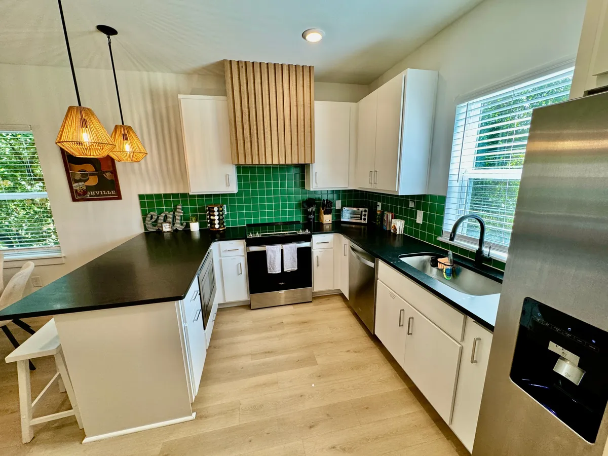 A bright, welcoming kitchen featuring a striking green tile backsplash, a sleek black countertop, a wooden range hood, and a central kitchen island. The space is well-lit and designed for both cooking and socializing, with modern appliances visible and plenty of counter space for meal prep.