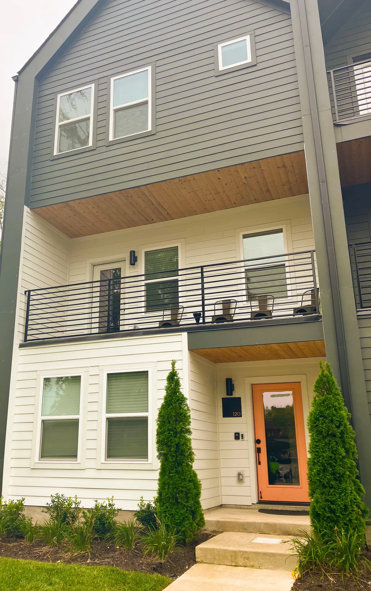 An exterior shot showing a welcoming orange door and a balcony furnished with chairs, framed by lush greenery and tall shrubs. The setting is perfect for relaxing with morning coffee or catching up with friends at sunset.