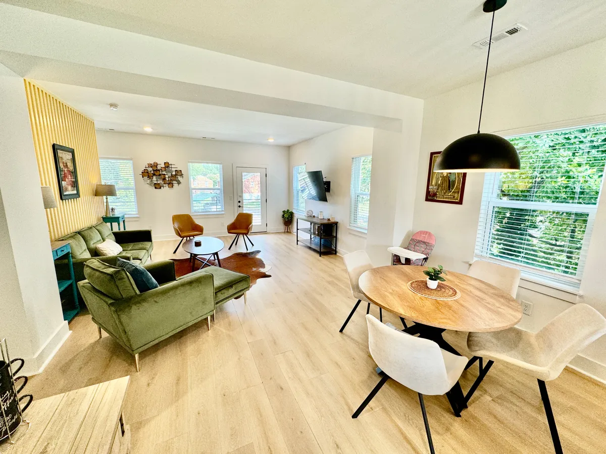 A cozy dining area adjacent to the kitchen, featuring a green sofa, a round wooden dining table, decorative wall art, and large windows. The space is set up for family gatherings, casual dining, and entertaining guests, with natural light creating a warm, connected atmosphere.