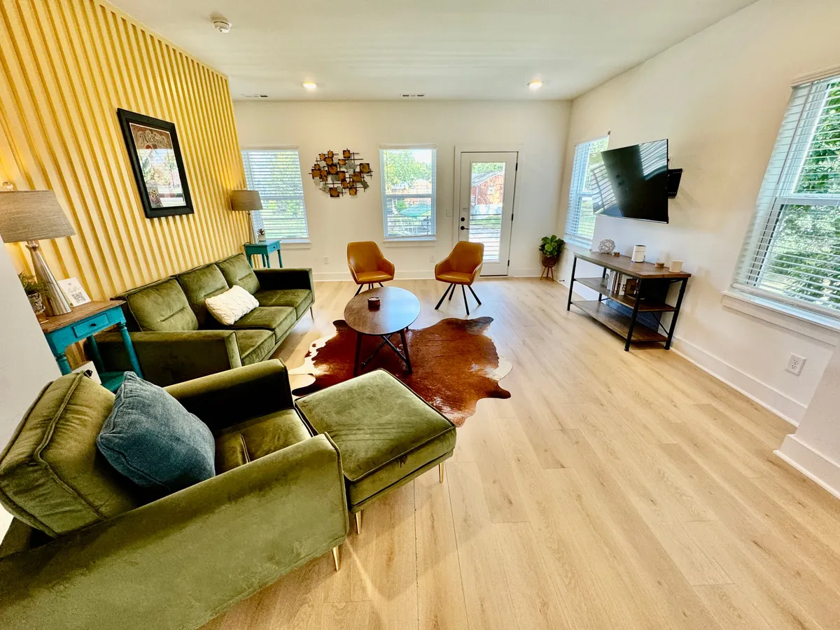 A cozy living room anchored by a green sofa and orange accent chairs, surrounding a wooden coffee table. Decorative wall art adds personality, and large windows flood the space with natural light. The space is open, connecting visually with the dining area, and is perfect for both relaxation and gathering.