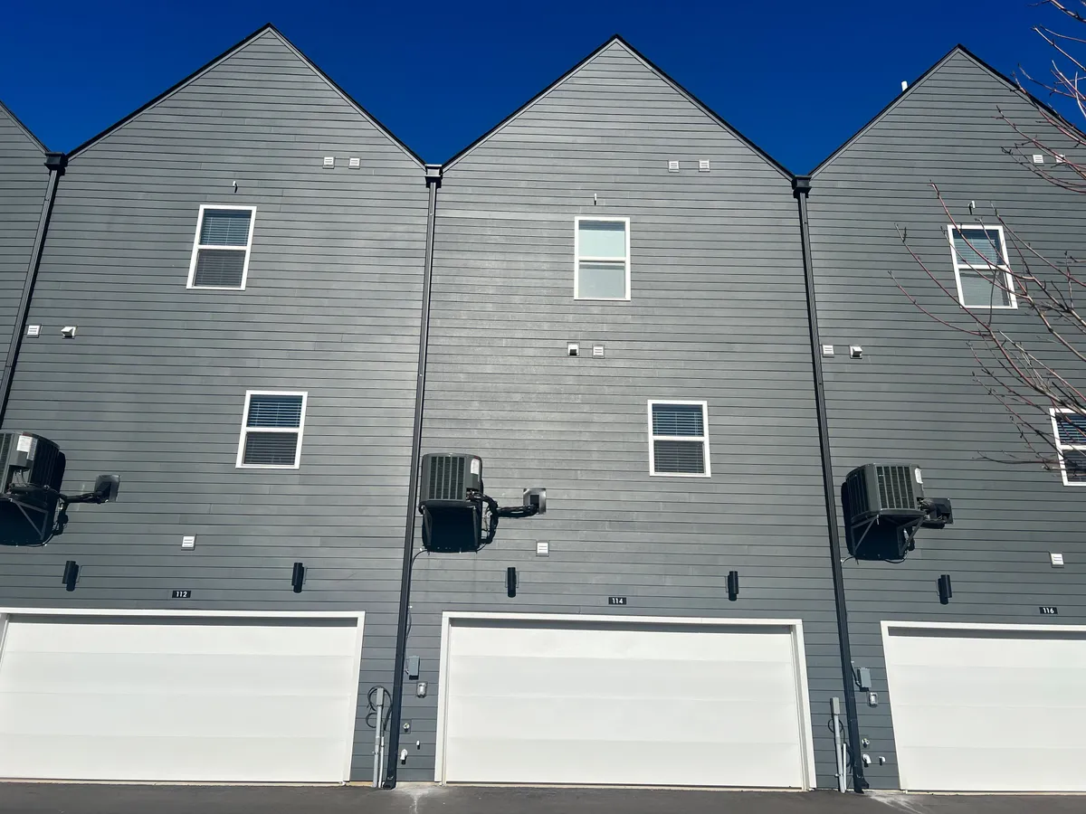 A view of modern townhomes with gray siding and white garage doors, set in a welcoming, residential community. The broad driveway and open environment hint at spacious outdoor areas ideal for walking dogs and casual strolls.